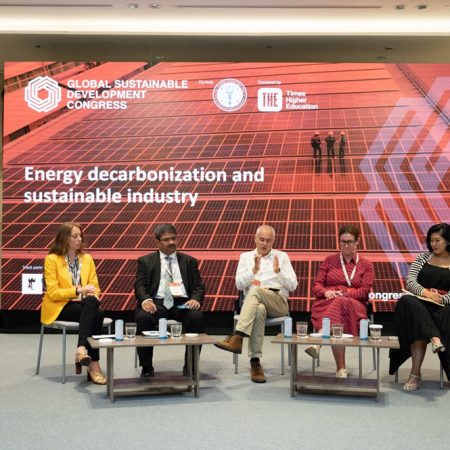 Five members of a conference panel sitting on a stage having a discussion, with a large promotional banner behind them