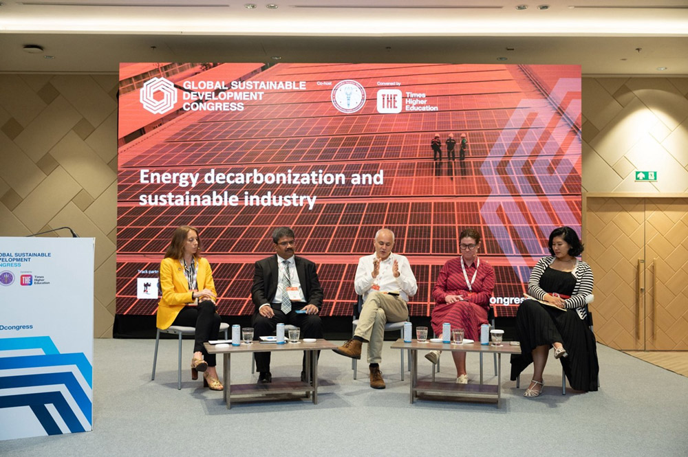 Five members of a conference panel sitting on a stage having a discussion, with a large promotional banner behind them